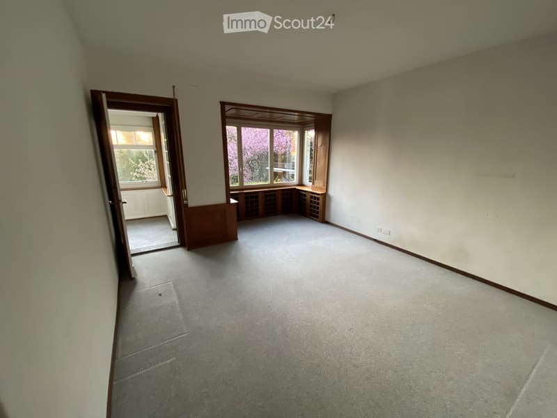 Empty room with light-colored walls, carpeted floor, large windows, and built-in wooden shelving unit.