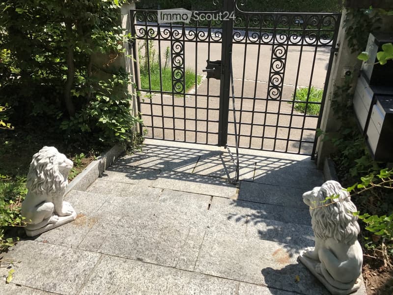 Garden entrance with a decorative wrought iron gate, flanked by two stone lion statues on a paved pathway.