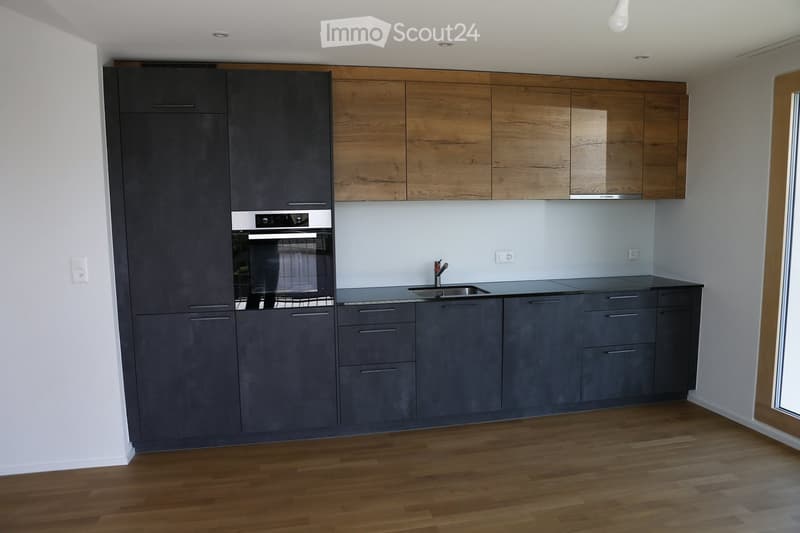 Modern kitchen with grey and wooden cabinets, a sink, and an oven. The floor is wooden, and the walls are white.
