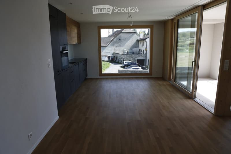 Empty room with wooden flooring, grey cabinets, and large windows overlooking a street and parked cars.
