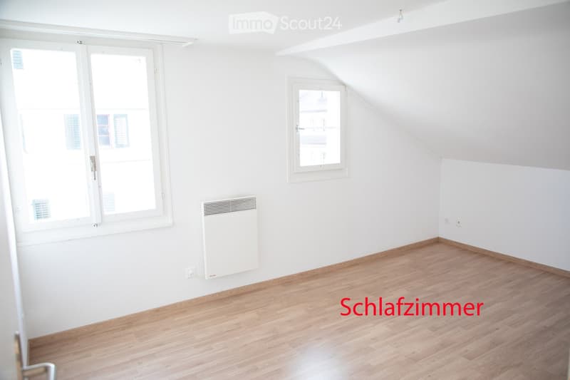Empty room with white walls, wooden flooring, and two windows, one with a radiator below it.