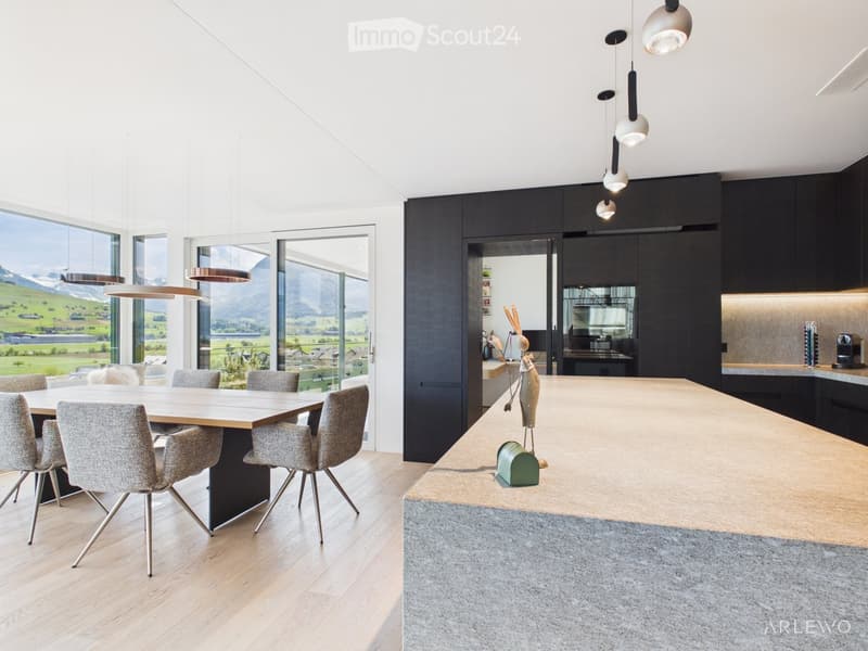 Modern kitchen and dining area with large windows offering a scenic mountain view, featuring a wooden dining table, grey chairs, a kitchen island, pendant lights, and sleek black cabinets.