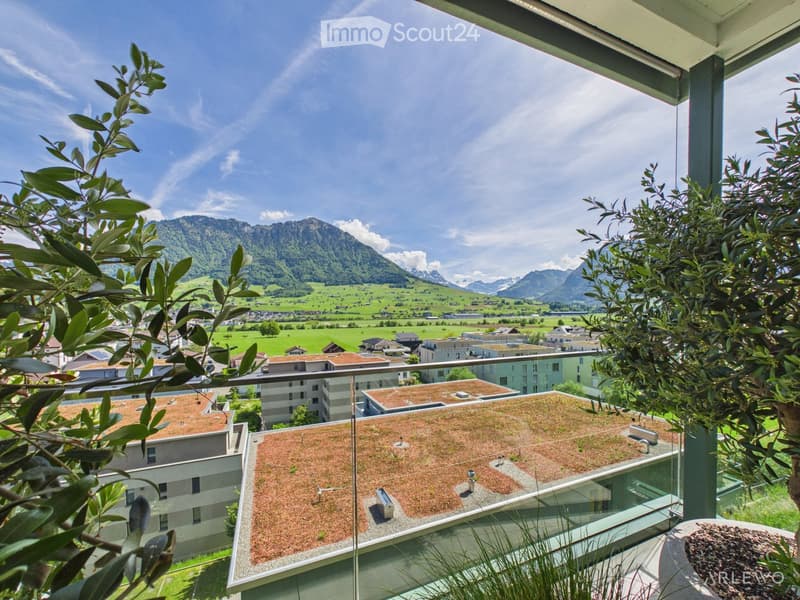 Balcony with a view of a green valley, mountains, and surrounding buildings. The balcony has a glass railing and is partially covered by plants.