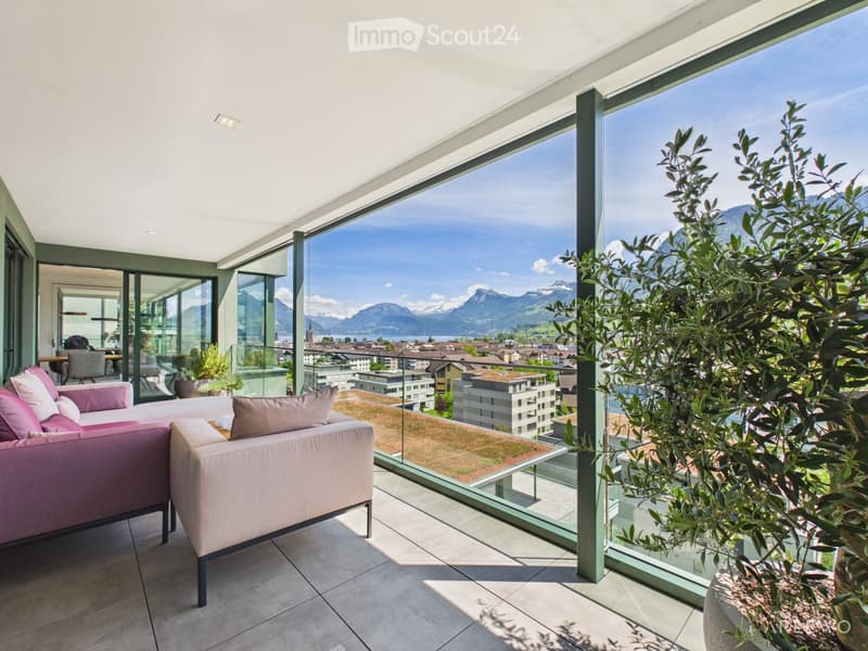Modern balcony with large glass windows offering a scenic mountain view, featuring a light pink sofa, a small table, and potted plants.