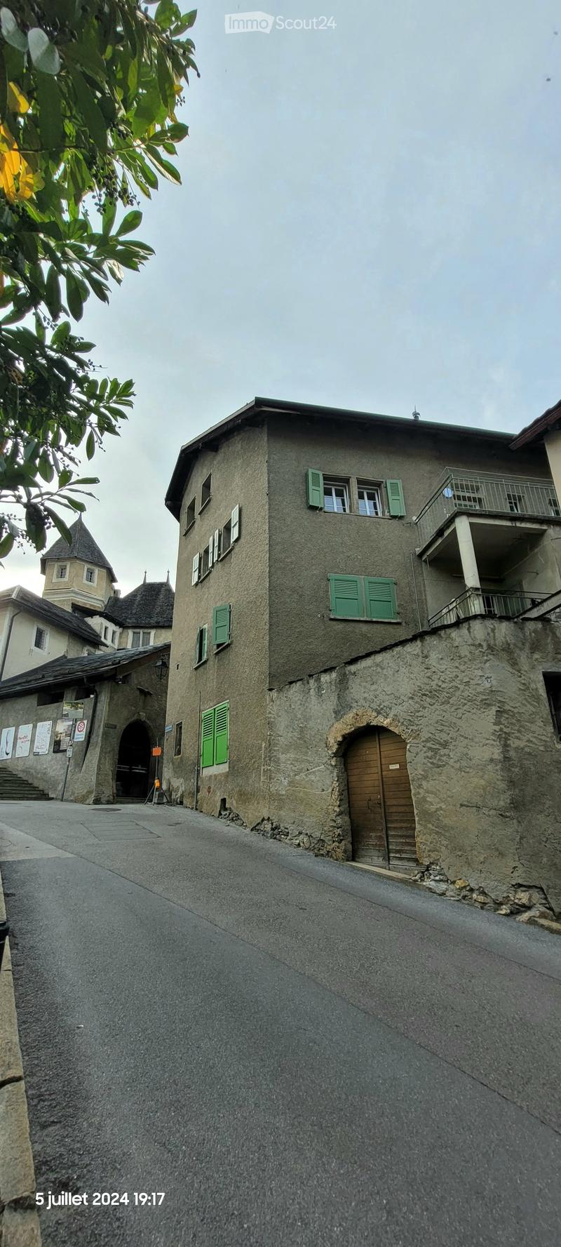 A multi-story stone building with green shutters and a balcony, located on a narrow street with a stone archway entrance.