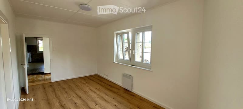 Empty room with wooden flooring, white walls, and a large window with white frames. A door leads to another room, and a radiator is visible on the wall.