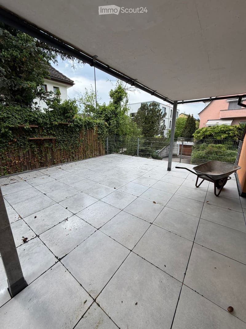 Empty patio with concrete tiles, metal railing, and a wheelbarrow in the corner. Surrounded by greenery and neighboring houses.