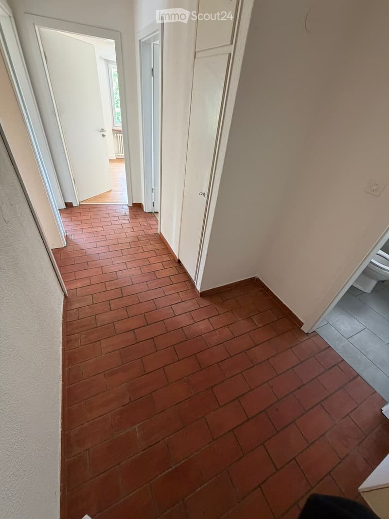 A narrow hallway with terracotta tiled flooring and white walls, featuring several open doors leading to adjacent rooms.