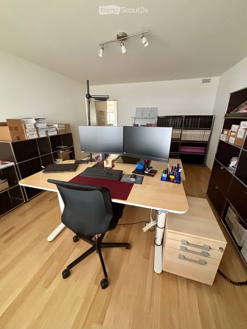 A modern, well-lit home office with a large wooden desk, ergonomic chair, dual monitors, laptop, keyboard, mouse, and various office supplies. Shelves with books and storage boxes are visible in the background.