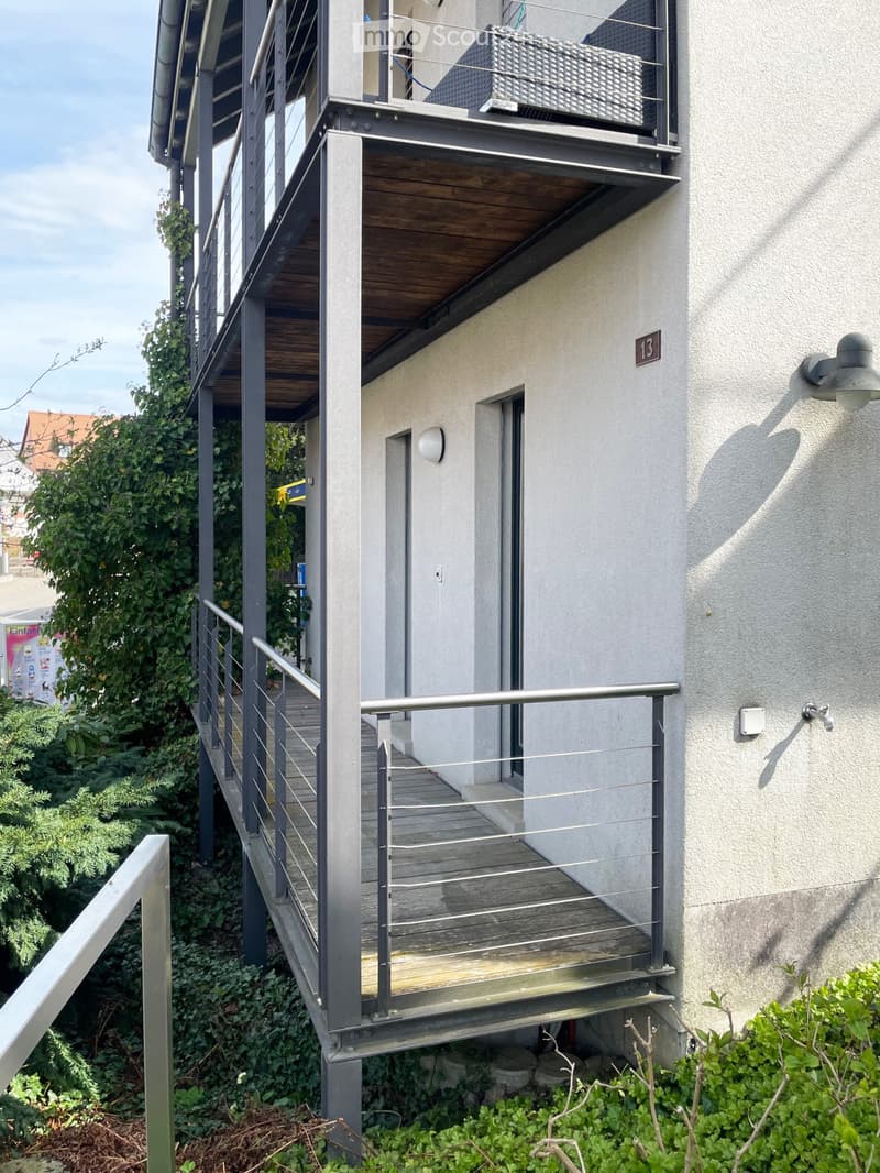 Balcony with wooden decking, metal railing, and glass panels, attached to a modern building with a light-colored facade.