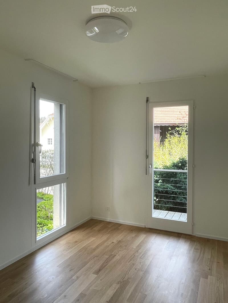Empty room with light green walls, wooden flooring, and large windows with sliding doors leading to a balcony or outdoor area.