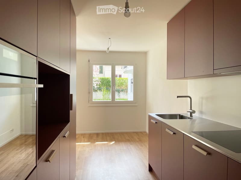 Modern kitchen with light grey cabinets, stainless steel sink, and wooden flooring. Features a window with a view of greenery outside.