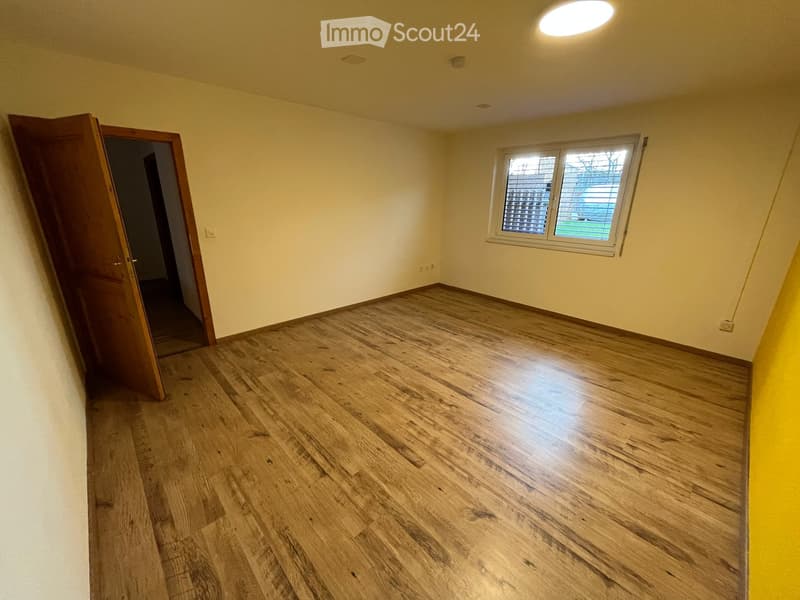 Empty room with wooden flooring, light yellow walls, and a single window with blinds. The room has a door leading to another space.