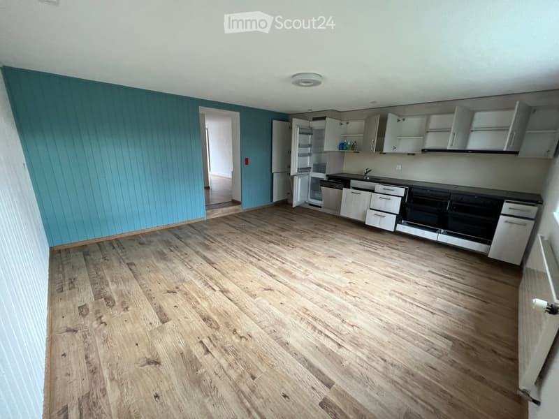 Empty kitchen with wooden flooring, white cabinets, and stainless steel appliances including a refrigerator, dishwasher, and oven. The walls are painted in a light blue color, and the space is well-lit with a ceiling light fixture.