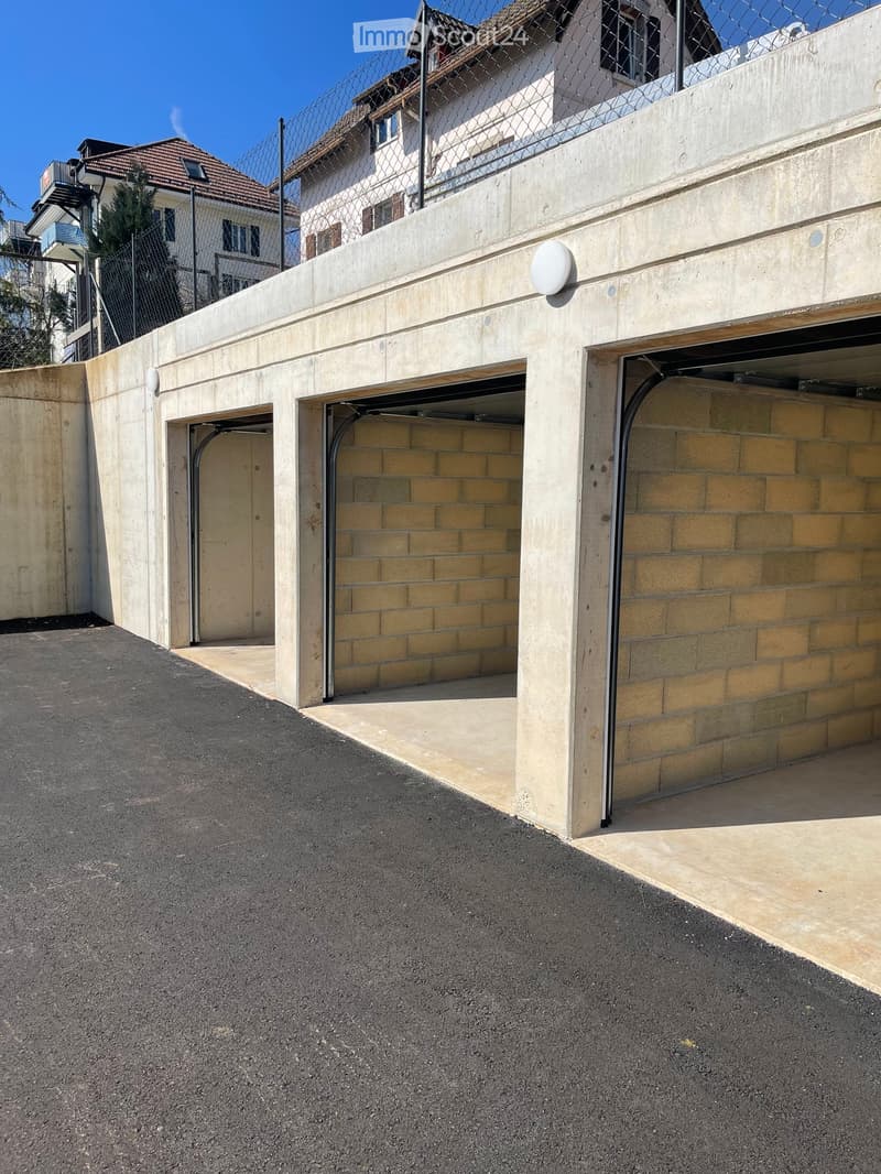 A modern concrete garage with multiple open doors, situated next to a fenced area and a residential building with a tiled roof.