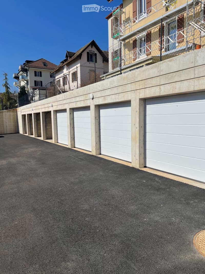 A row of white garage doors attached to a concrete wall, with a paved asphalt driveway in front and residential buildings in the background.