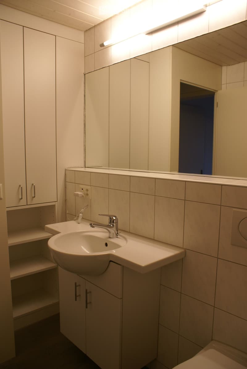 Bathroom with white tiles, sink, mirror, and cabinets. Power outlet and light fixture visible.