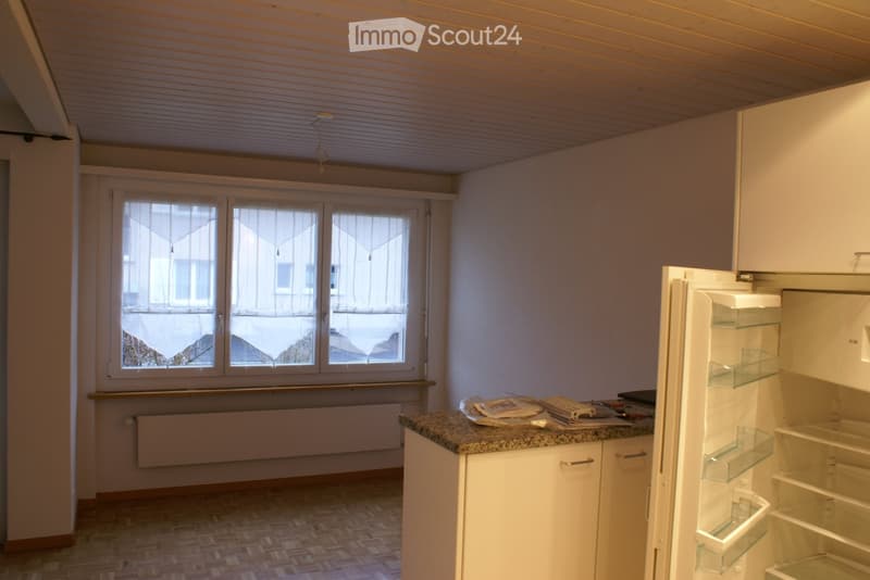 Empty room with white walls, wooden floor, and a window with curtains. A refrigerator is open, and there is a countertop with some items on it.