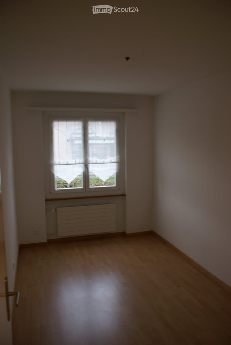 Empty room with white walls, wooden floor, and a window with white curtains. A white radiator is installed below the window.