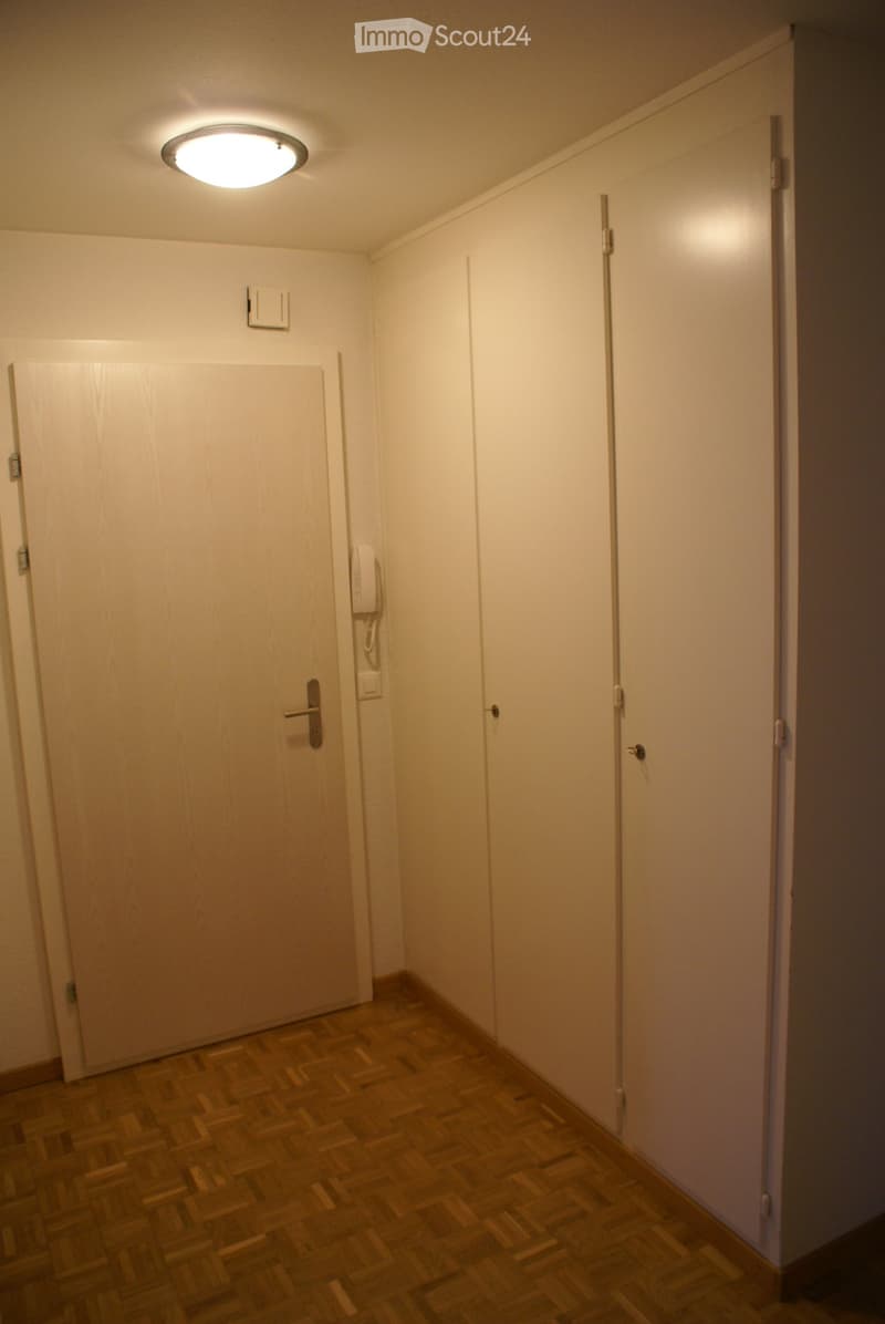An empty room with a white door, white walls, and wooden floor. There are two white cabinets on the right side of the room, and a telephone is mounted on the wall next to the door.