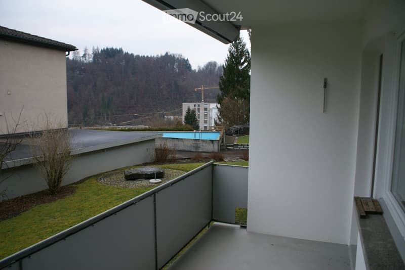 A balcony with a metal railing, overlooking a grassy area with a rock and a swimming pool in the distance.