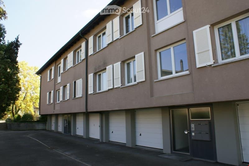 A three-story building with multiple windows and white shutters. The building has a parking area in front with several garages.