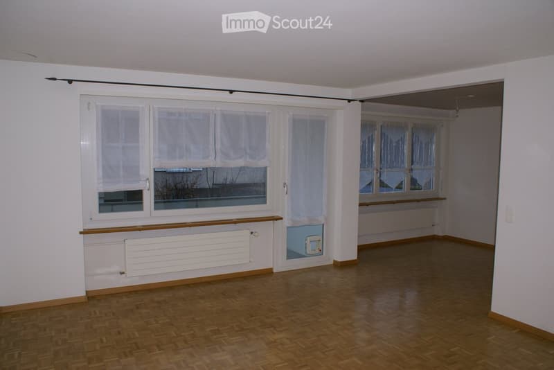 Empty room with white walls, wooden floor, and large windows with white curtains. There are white radiators on the walls and a door leading to another room.
