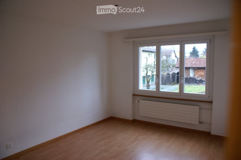 Empty room with white walls, wooden floor, and a window with a view of a house and trees outside.