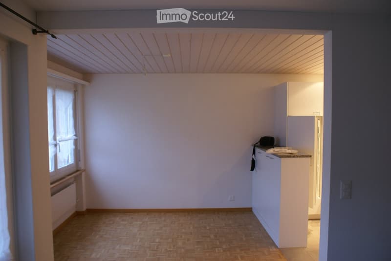Empty room with white walls, wooden floor, and a window with white curtains. A white cabinet with a countertop is on the right side.