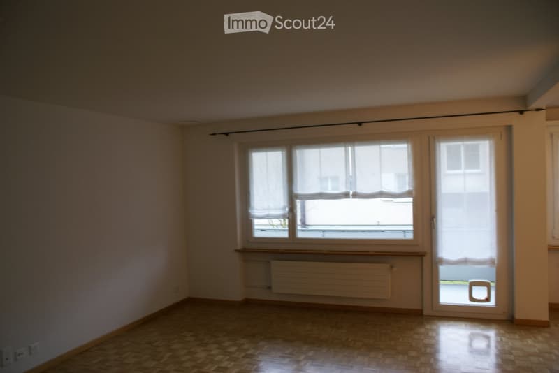 Empty room with white walls, wooden floor, and a window with white curtains. There is a radiator below the window and a door leading to another room.