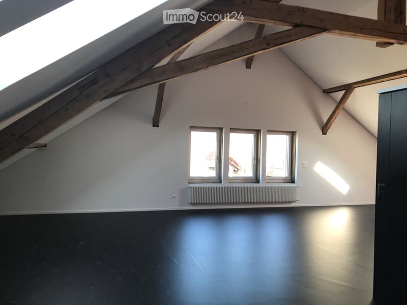 Empty attic space with wooden beams, three windows, radiator, and dark flooring.