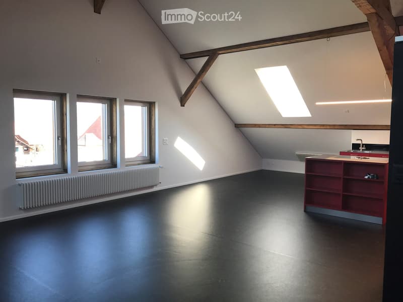 An empty attic room with exposed wooden beams, large windows, and a skylight, featuring a dark floor and white walls.