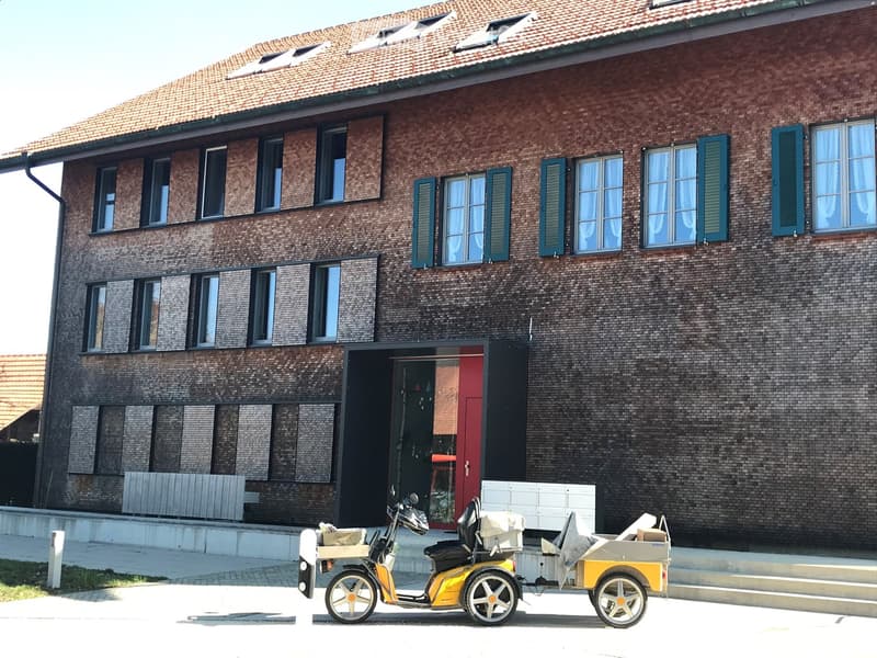 Two-story brick building with multiple windows and a red-tiled roof, featuring a raised ground floor entrance and a small yellow vehicle parked in front.