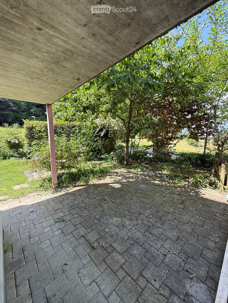 A covered outdoor area with a stone-paved floor, surrounded by lush greenery and trees, with a concrete roof supported by a pink column.