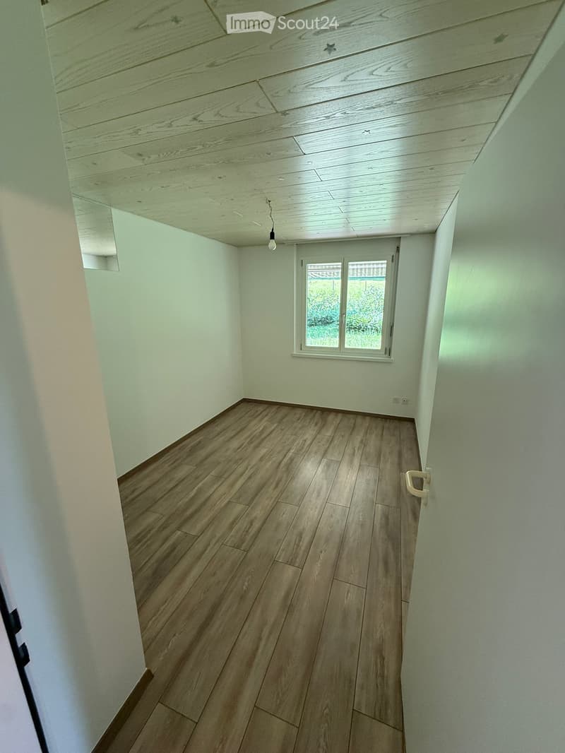 Empty room with wooden flooring, white walls, and a window with a view of greenery outside. The ceiling features exposed wooden beams and recessed lighting.