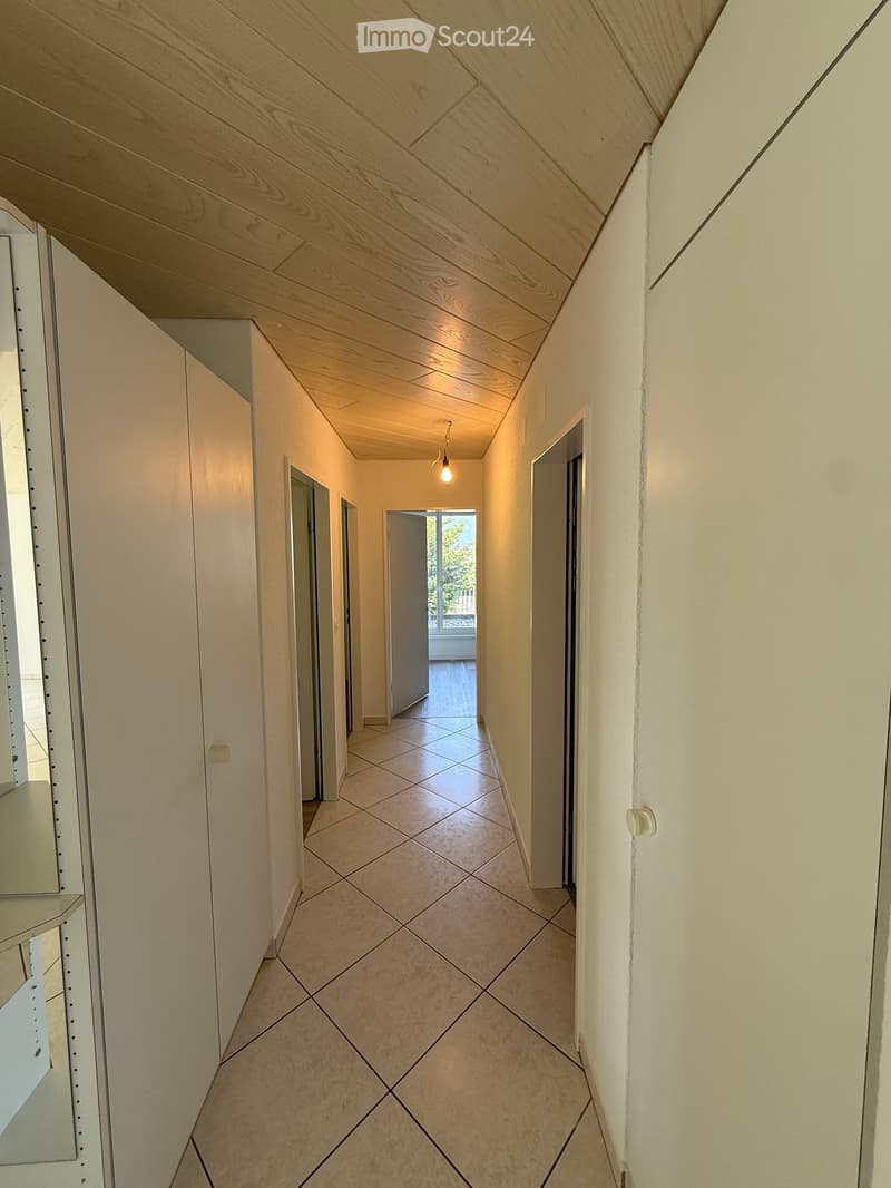 A narrow hallway with light-colored walls, tiled floor, and a single hanging light fixture. The hallway leads to an open doorway showing a glimpse of an outdoor area with a view of greenery.
