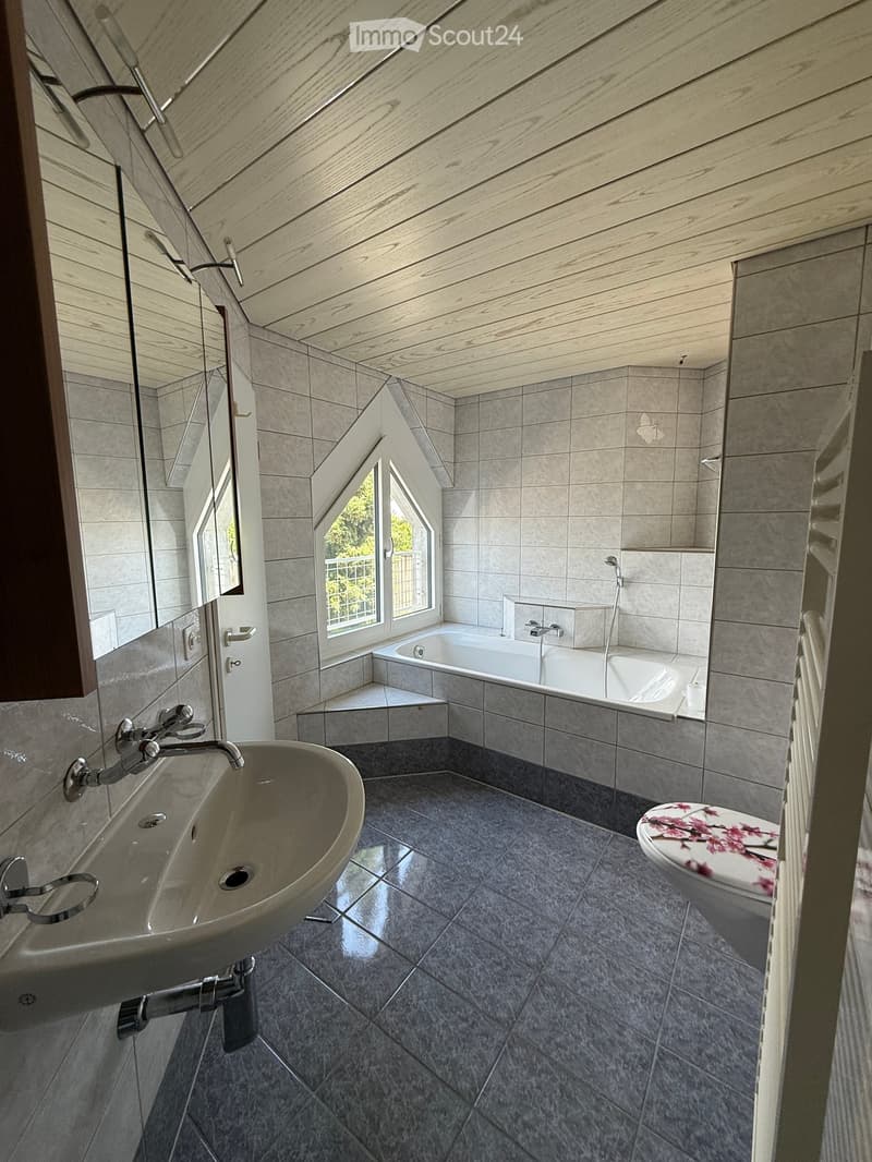 Modern bathroom with grey tiles, a sink, bathtub, toilet, and a large window letting in natural light.