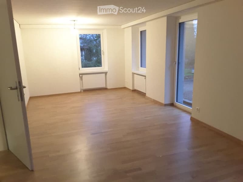 Empty room with wooden flooring, white walls, and large windows, including a sliding glass door leading outside.