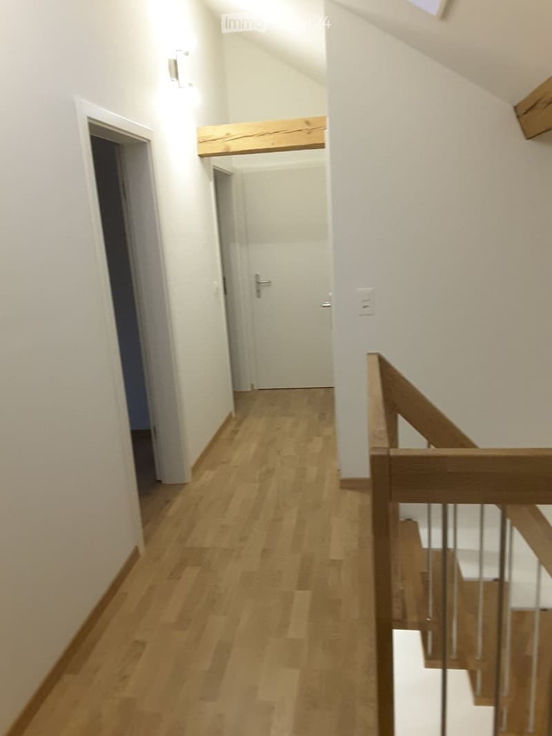 A narrow hallway with light wooden flooring, white walls, and a wooden staircase railing on the right side. The hallway leads to a closed door at the end.