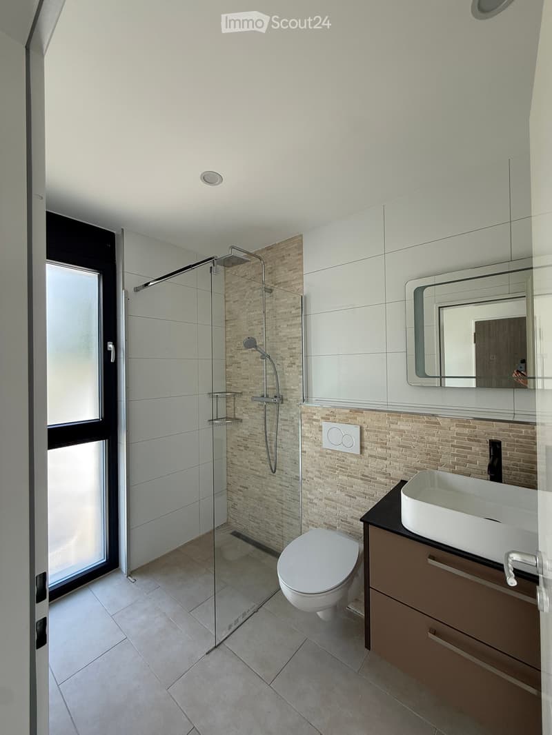Modern bathroom with a glass-enclosed shower, toilet, sink, and mirror. Features beige brick accents and white tiles.