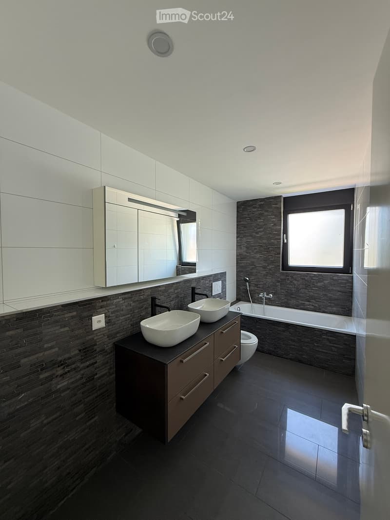 Modern bathroom with dark tiled walls and floor, double sink vanity with two modern sinks, a bathtub, and a large mirror above the sink.