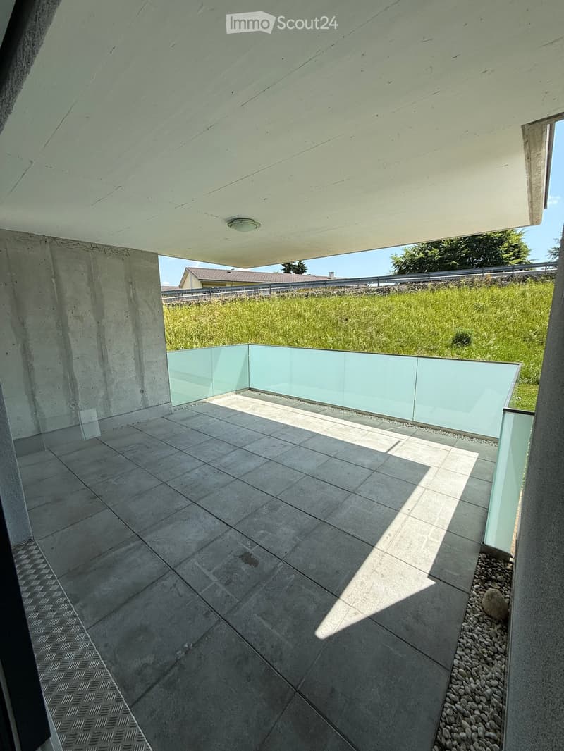 Modern concrete balcony with tiled floor, glass railing, and a view of a grassy area.