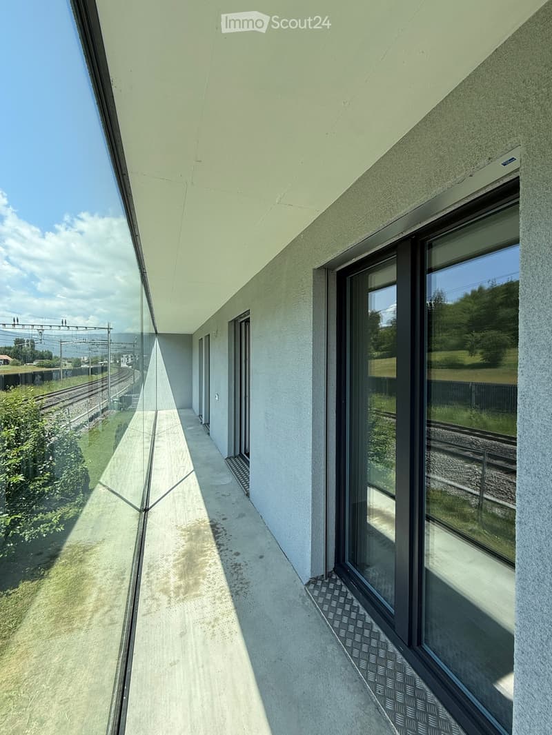Modern balcony with large glass railings, concrete floor, and views of railway tracks and greenery.