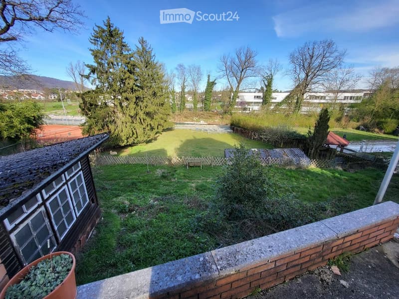 View from a balcony overlooking a grassy area with trees, a wooden fence, and a small shed. The background includes a tennis court and distant buildings.
