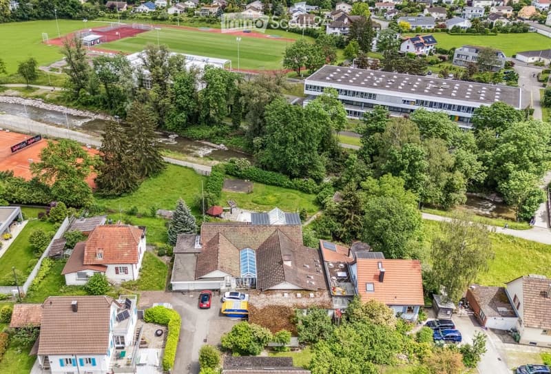 Aerial view of a residential area with multiple houses, a school building, sports fields, and a river running through the landscape.