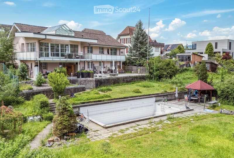 A two-story house with a modern design, featuring a large balcony, a swimming pool, and a well-maintained garden with various plants and outdoor furniture.