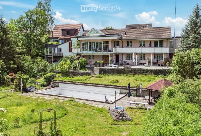 A two-story house with a modern design, featuring a large balcony, multiple windows, and a spacious garden with a swimming pool, patio furniture, and lush greenery.
