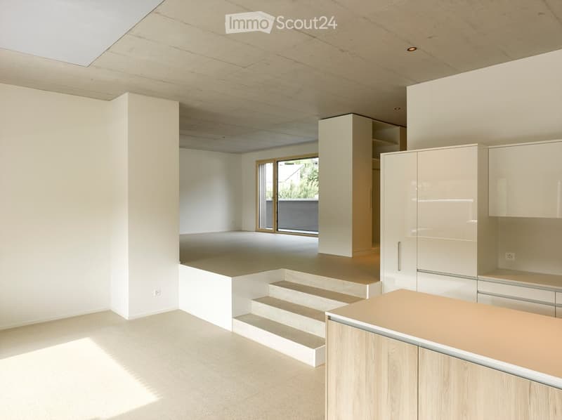 Modern, minimalist kitchen with white cabinetry, light wood accents, and a raised platform area. Features large windows and a skylight, with a clean, uncluttered design.