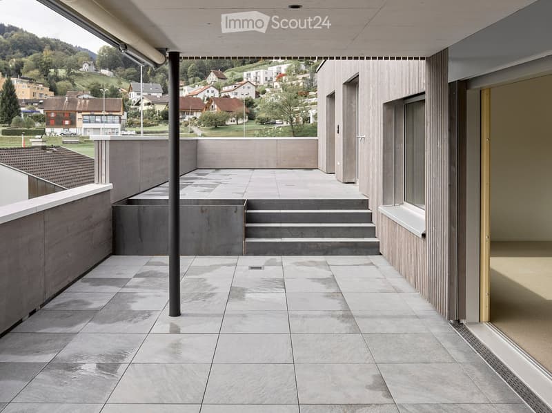 Modern balcony with concrete steps leading up, grey tiled floor, wooden railings, and a view of a residential area with houses and greenery.
