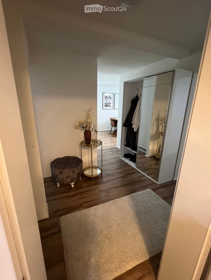 Furnished Room. A hallway with wooden flooring, a small round table with a vase, a plush stool, a white rug, and a mirrored wardrobe with clothes inside.